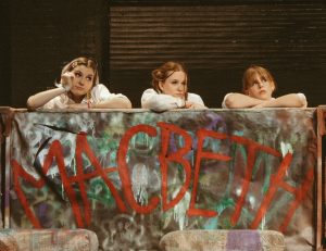 Three girls in school uniforms lean over the back of a couch covered with green, purple, white, and grey spray paint. MACBETH is written on the couch in red spray paint. 