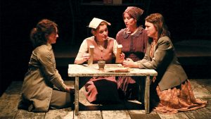 Four women in 1940's dresses and cardigans sit on their knees around a small wooden table. One woman is holding two pieces of paper with a small clay cup and burlap sack on the table in front of her.