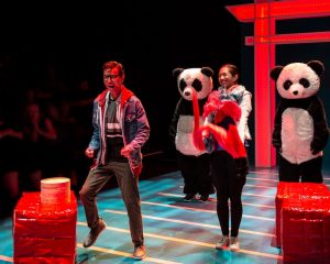 A man and a woman dance on stage with two people in panda suits and a red pagoda arch behind them.