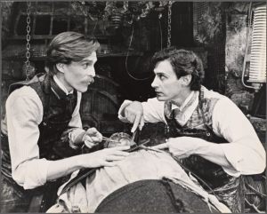 In a black and white photo, two men in white button up shirts, vests, and cravates talk to each other while holding scissors and tweezers over the heart of the monster laying on a table. In the background are chains, coils, and grates on the walls. 