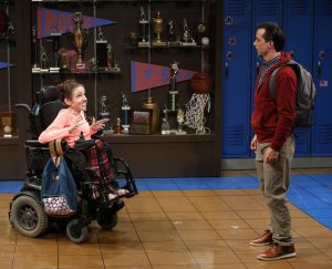 In a school hallway with lockers and full trophy case, a girl with a pink shirt in an electric wheelchair is talking to a boy standing with a backpack.