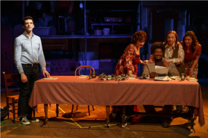 Shows for Days production photo. A long dining room. To the left, a man stands and looks up into a spotlight. To the right, a group of 3 women surround a man who is holding a letter and smiling.